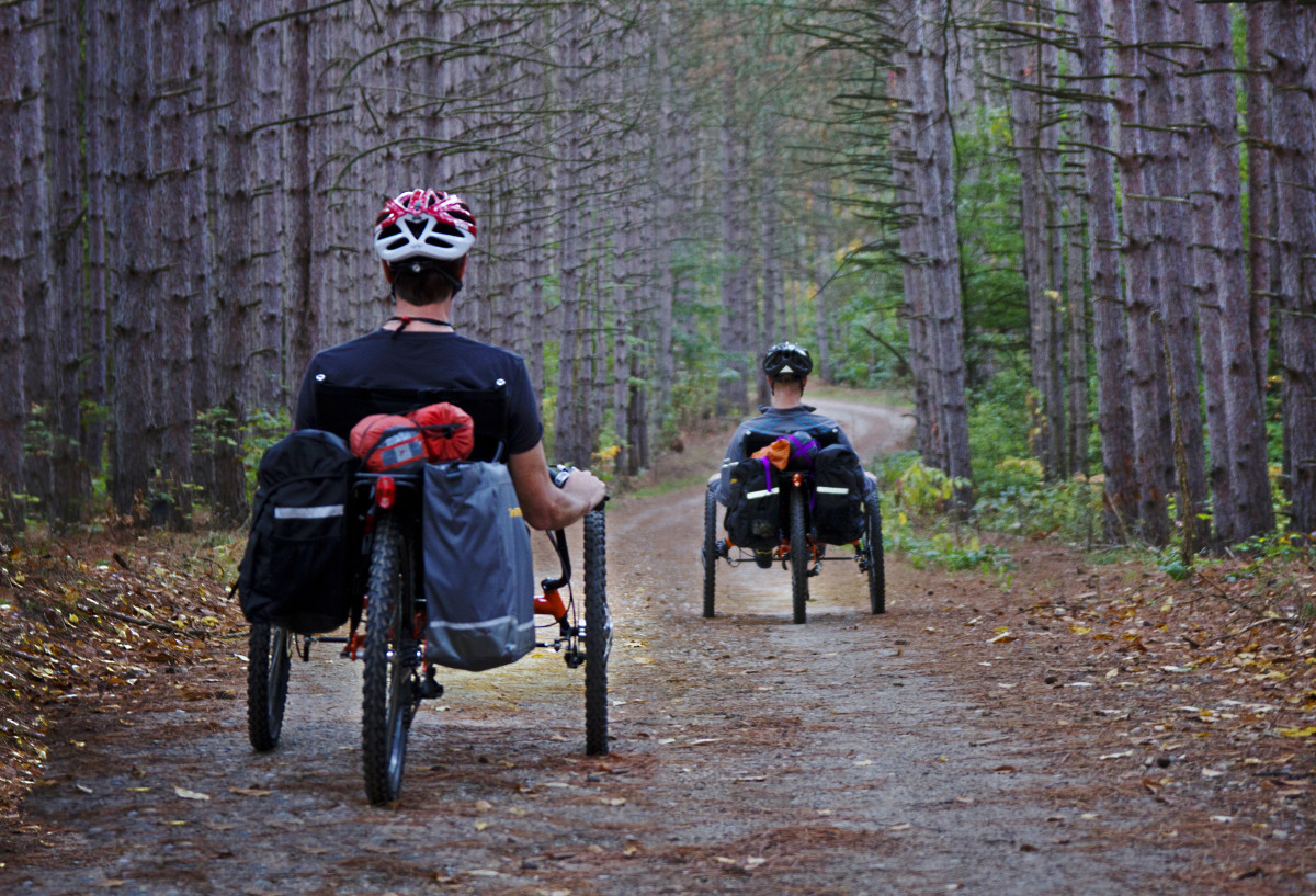 Two men ride their recumbents through a forest path, both with camping gear strapped to the backs of their bikes