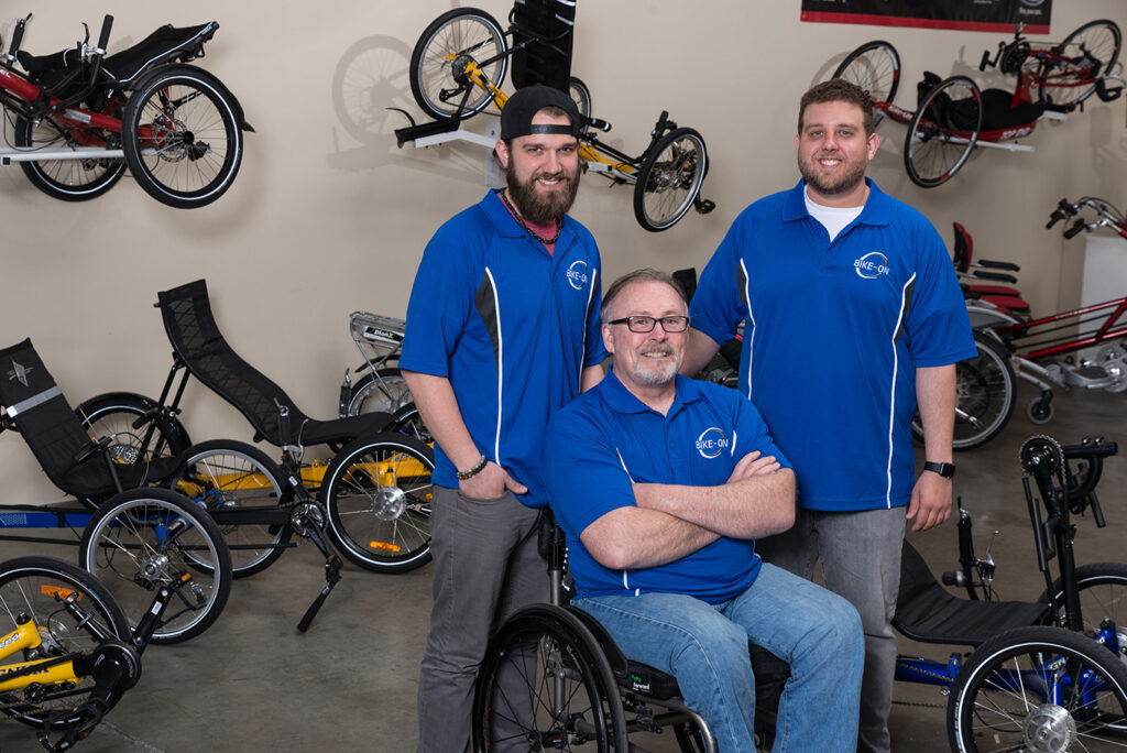 three men wearing blue polo shirts with recumbent bikes behind them. chris the general manager/ lead mechanic stands next to dan, ceo and scott is in front of them seated in his wheelchair the founder of bike ont