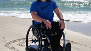 Man wearing sunglasses staring into camera on the sand in front of the ocean, he is riding a wheelchair with the Freedom Trax Off-Road attachment fixed to the bottom