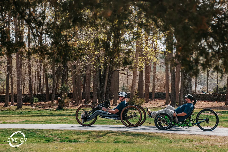 Two men ride along a park path, one in a Crosswind Handcycle, the only in a recumbent trike