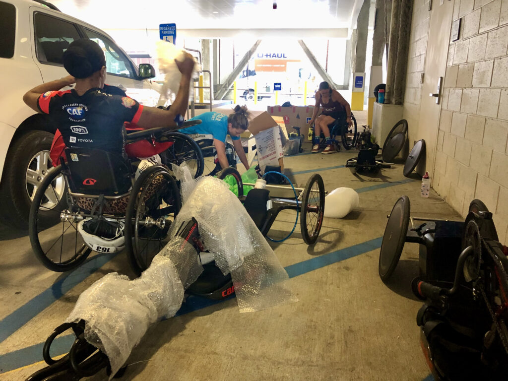 3 people in wheelchairs work on wrapping their handcycles in bubble wrap inside of a parking garage