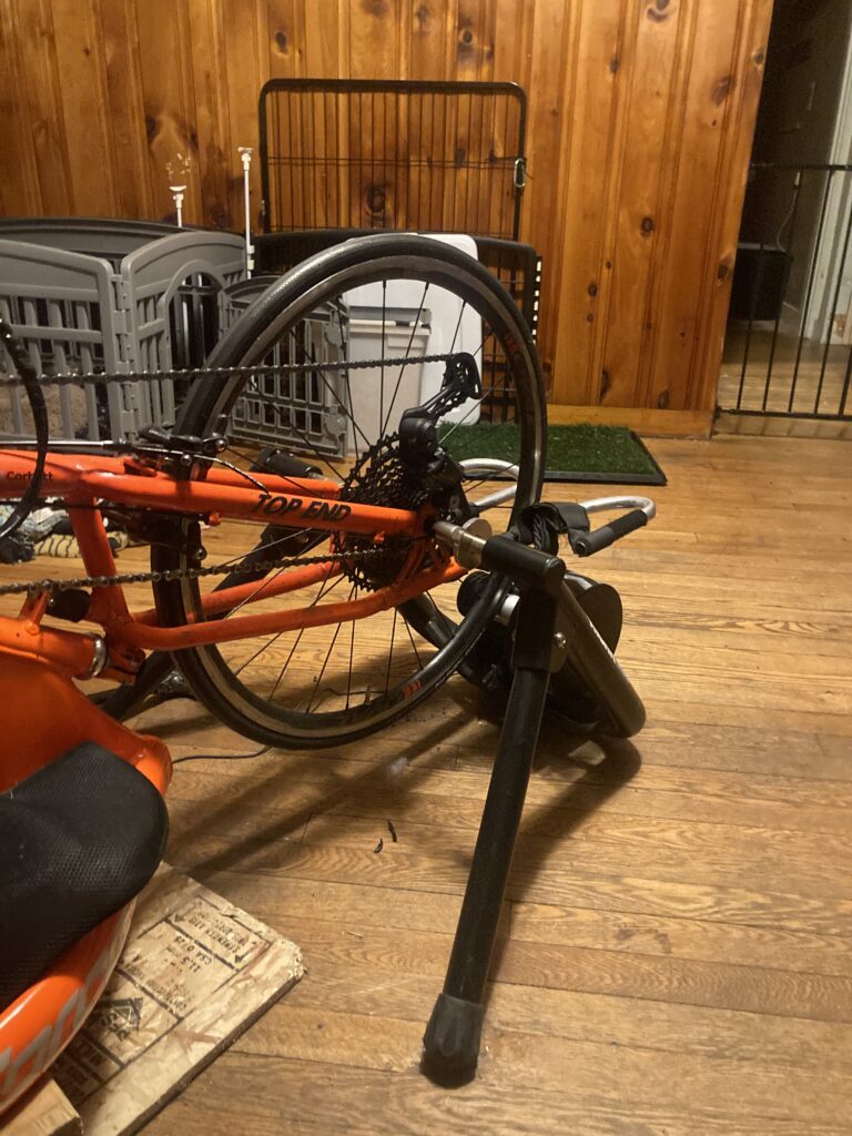The front wheel of a orange handcycle bike connected to a black stand, the Wahoo Kickr trainer. 
